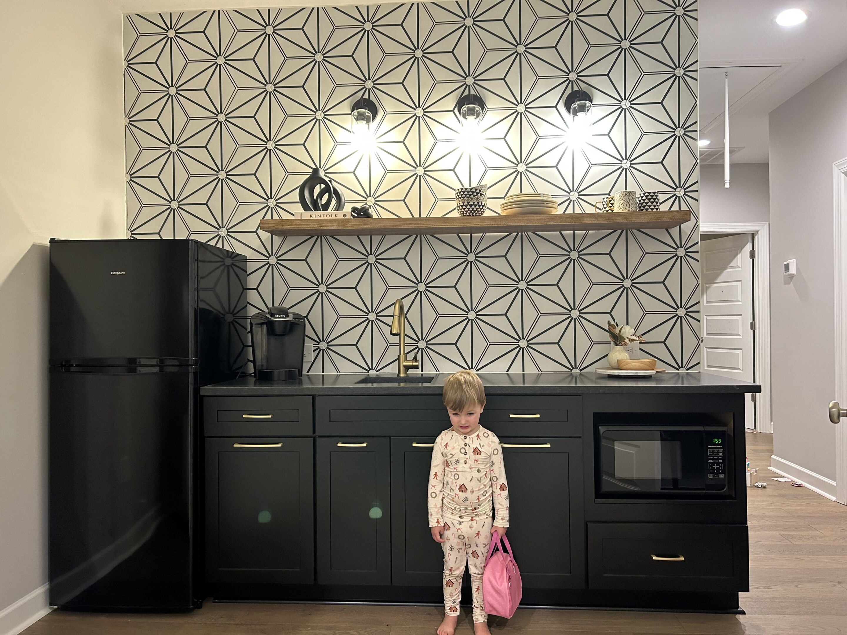 A little girl standing in the kitchen with her pink bag.